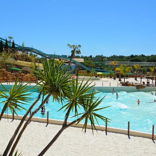 Un parque acuático con personas nadando en una piscina de olas, rodeado de palmeras y toboganes de agua bajo un cielo azul claro.