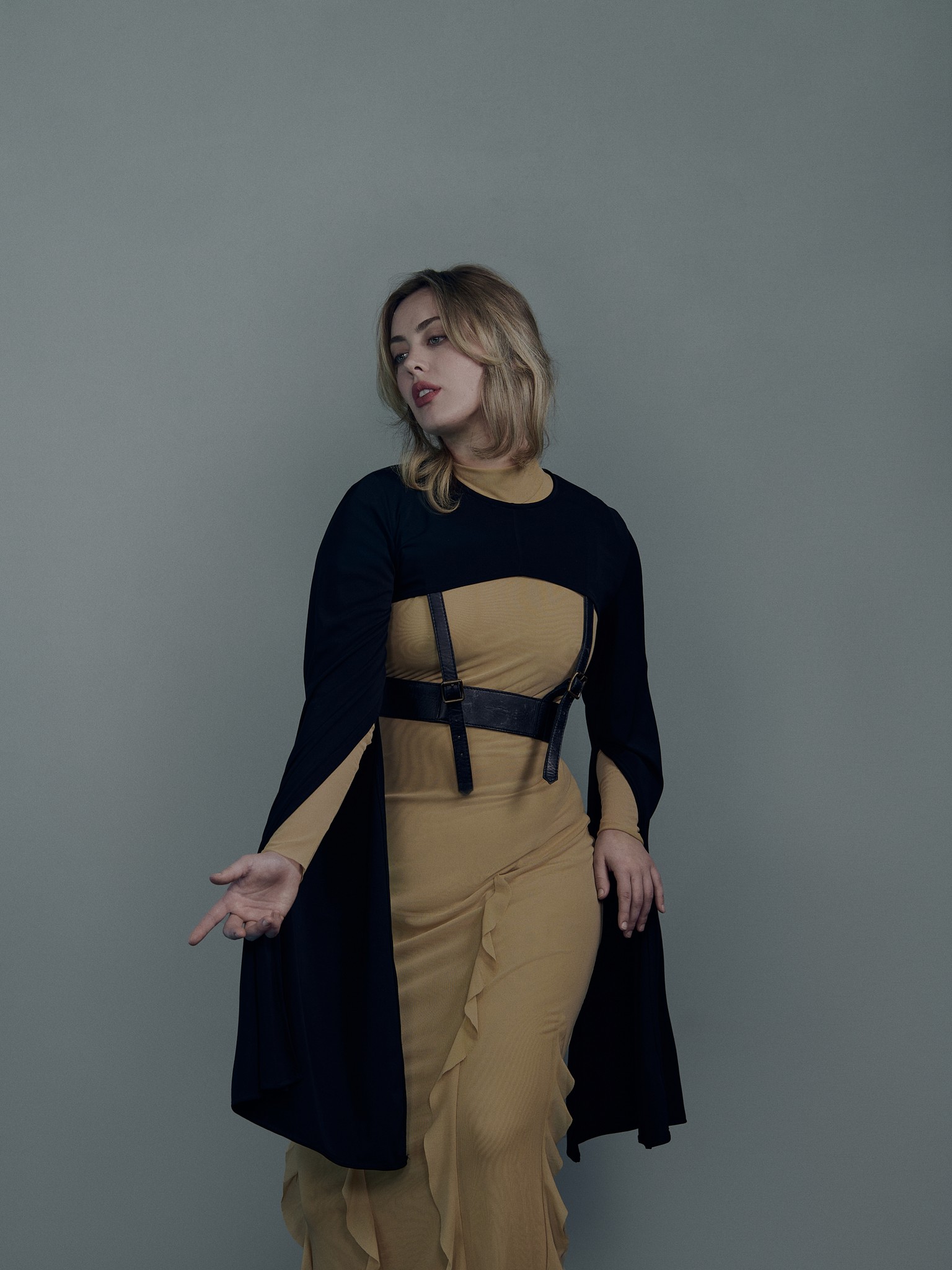 An editorial studio portrait of Madison Randolph in a modern classical pose. The photograph has a moody dramatic tone and expression. She is wearing a leather harness and cape. 