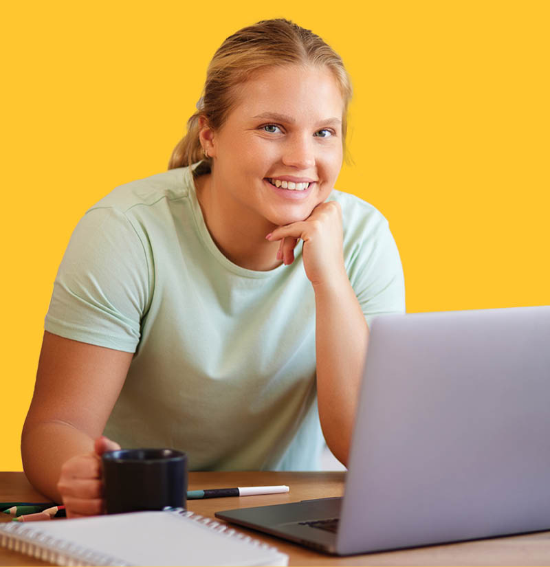 Adult learner sitting at a desk with a laptop, notebook, and coffee mug, looking toward the screen, against a yellow background.