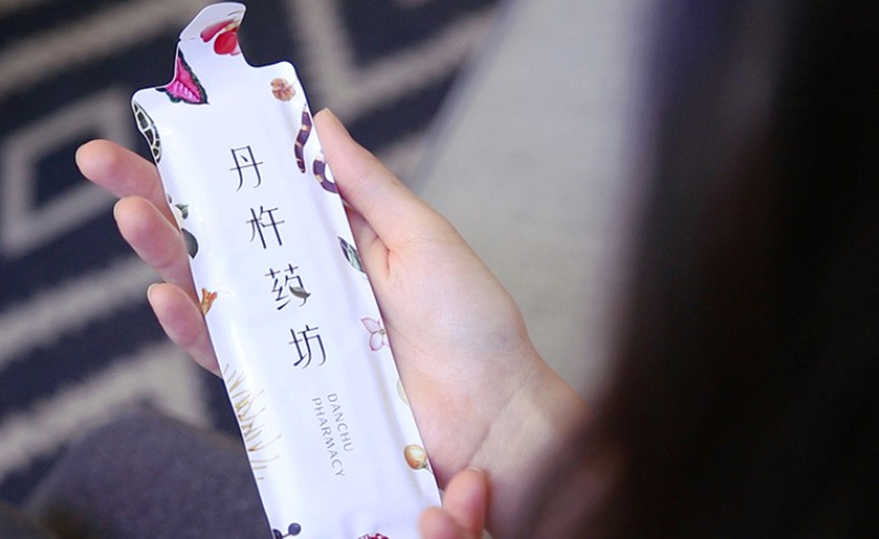 A hand holds a partially unwrapped white rectangular package. The package features Chinese characters and images of berries and leaves. The text "DANCHU PHARMACY" is printed on the lower portion of the package. The background is blurred, showing a patterned surface.
