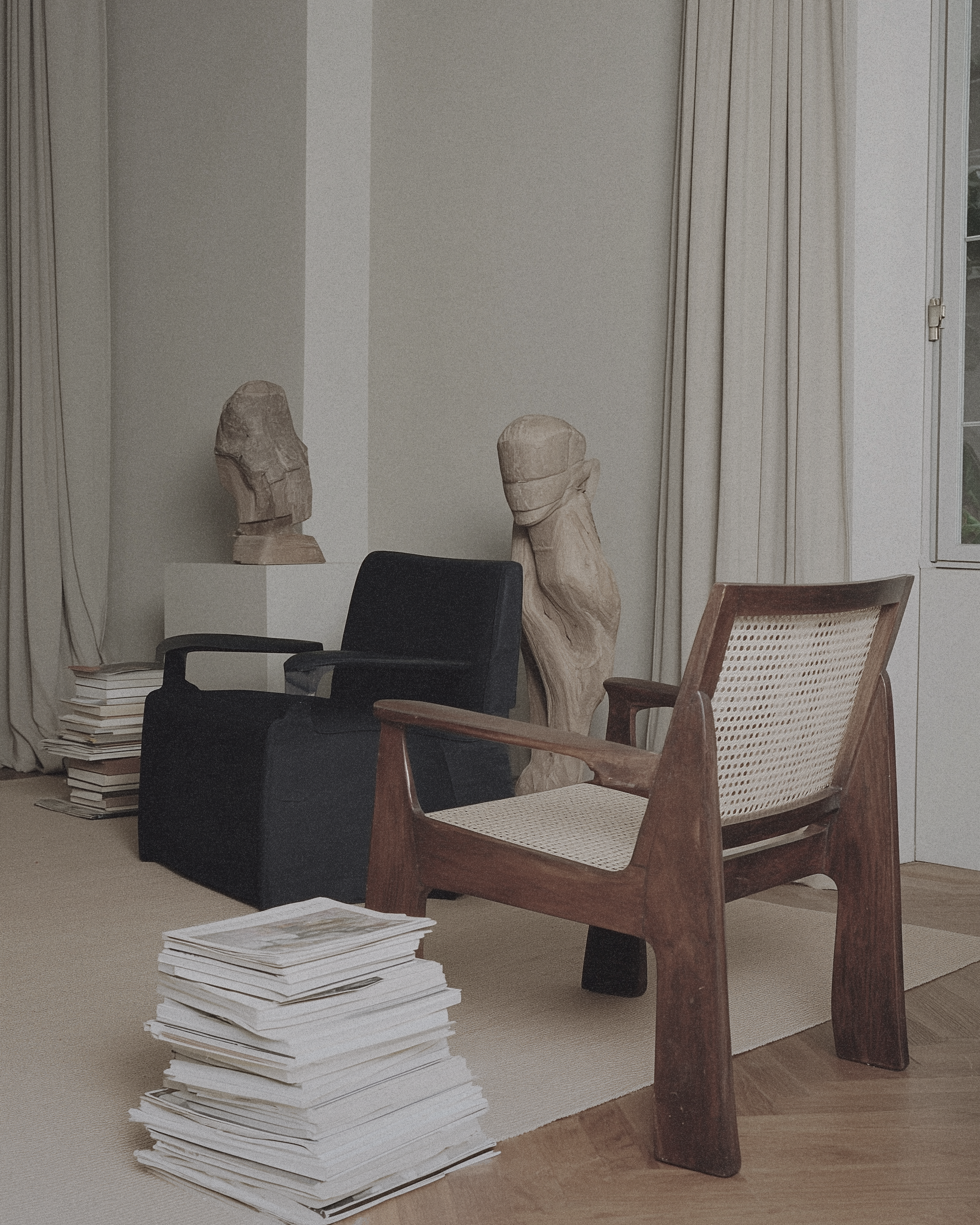 Neutral living room interior with a cane chair, books stacked on the floor, and sculptural decor