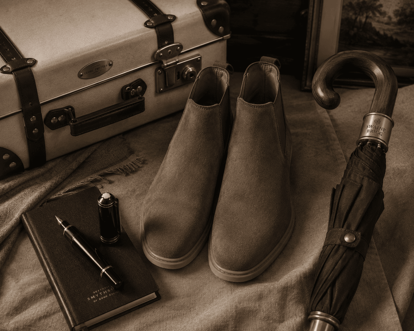 A refined travel flat lay featuring a pair of taupe suede Chelsea boots positioned on a neutral linen cloth. In the background sits a cream Globe-Trotter vintage-style suitcase with brown leather trim and brass hardware. To the right rests a Swaine Brigg umbrella with a polished wooden handle, and to the lower left, a Montblanc fountain pen lies beside a dark Smythson leather notebook.