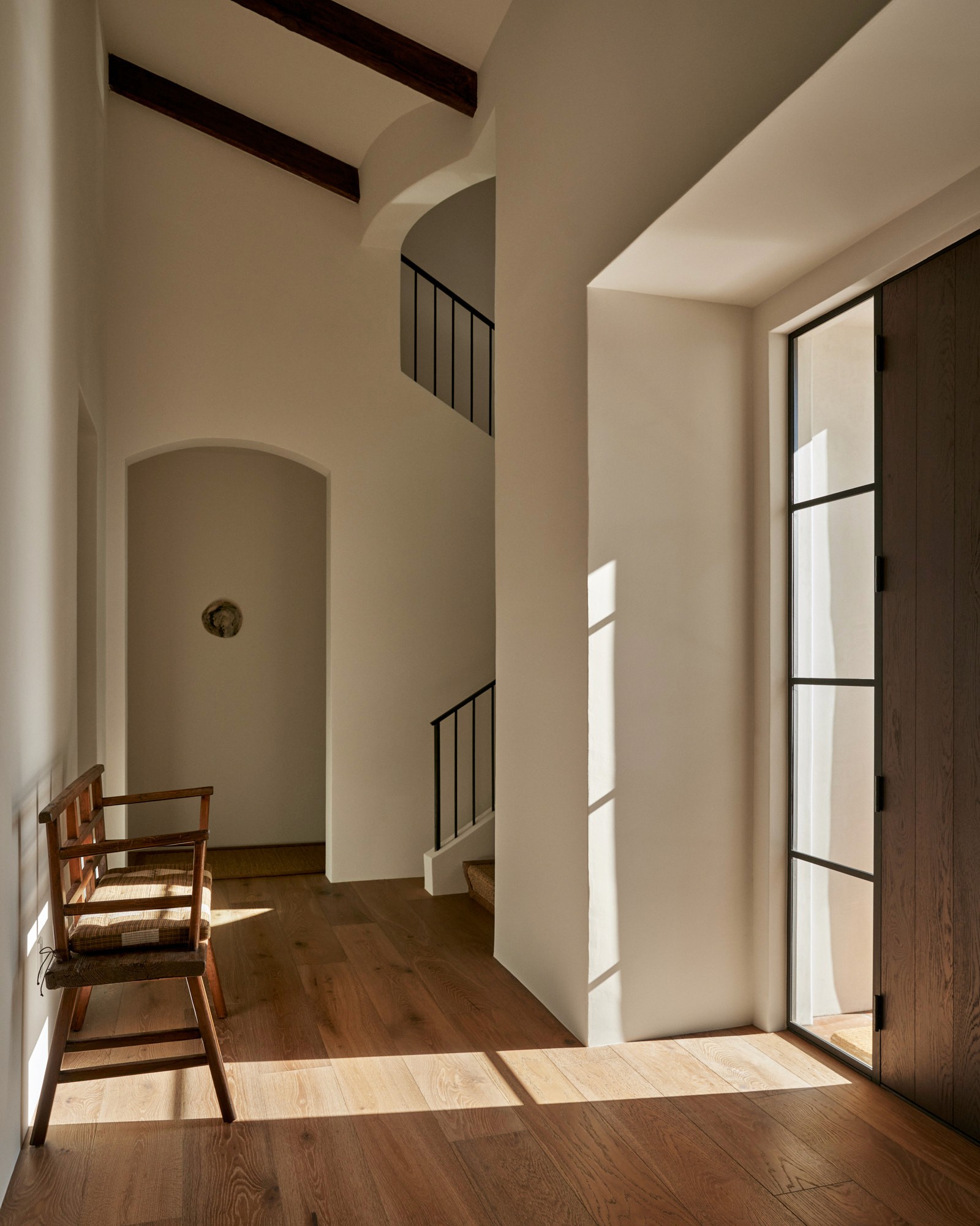 A double-height entry hall with white plaster walls, wide-plank oak floors, and a curving staircase with a wrought-iron railing. A single rush-seat chair sits in the natural light cast through a steel-frame door. A small, round artwork is set into the wall above the arched alcove.