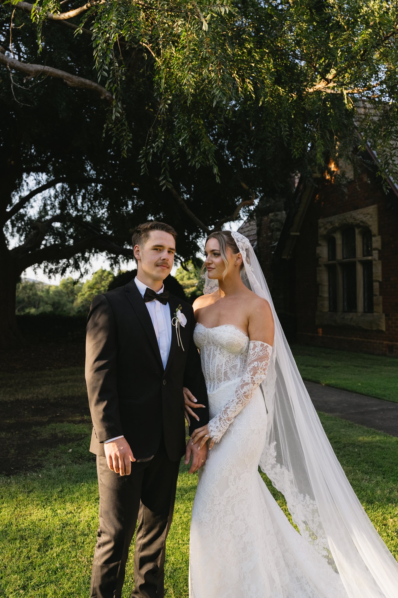 Bride in lace gown and veil with groom in black tuxedo stand under a sprawling tree beside a brick venue.