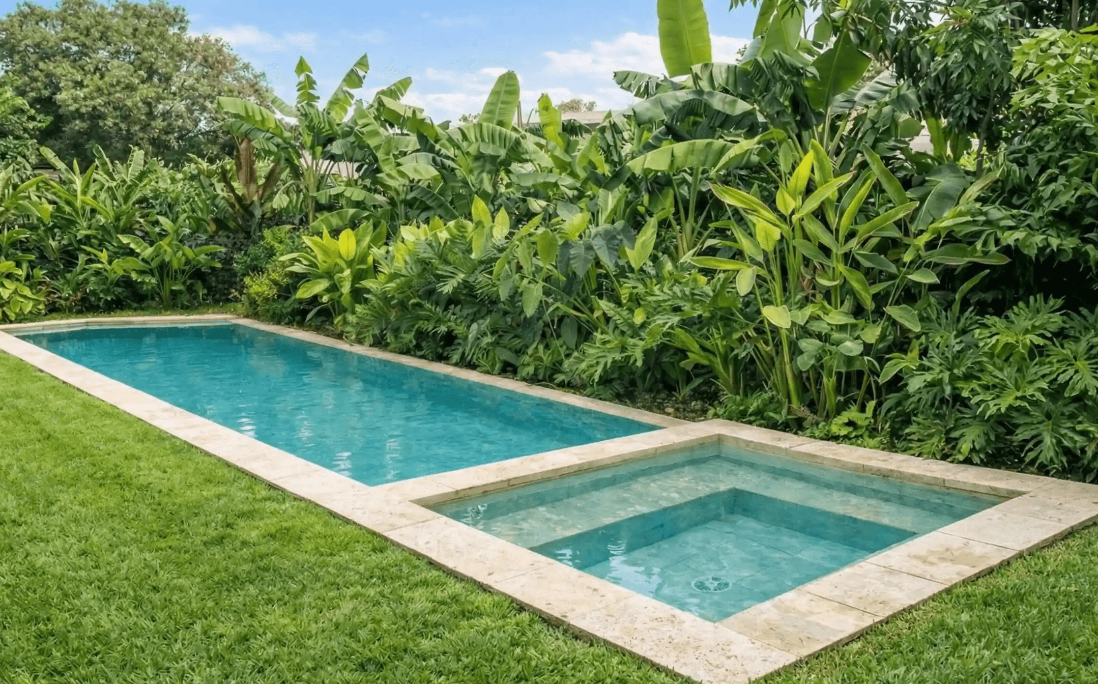 Piscina rectangular revestida en tonos claros con anexo para niños, rodeada de un exuberante jardín tropical con palmeras.