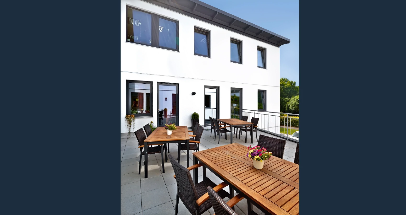 Terrasse mit mehreren Holztischen und Stühlen vor einem modernen weißen Gebäude mit großen Fenstern.