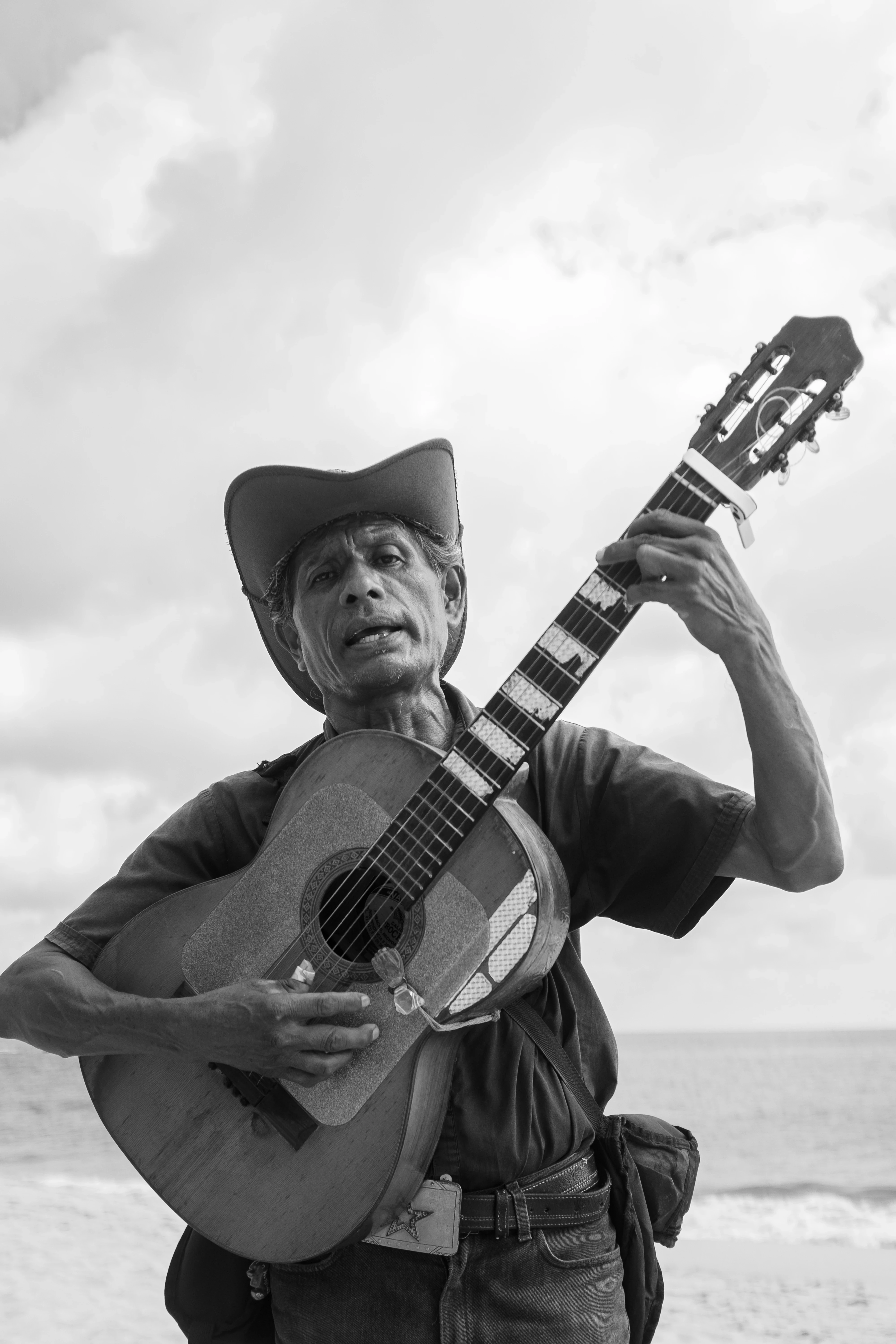 A grayscale image of a person wearing a cowboy hat, passionately playing an acoustic guitar at the beach, with a cloudy sky and ocean in the background.
