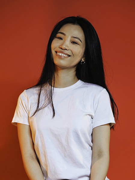 A young woman with long black hair stands against a solid orange-red background. She wears a plain white t-shirt and smiles directly at the camera. Her head is turned slightly to the right, and she has dark eyes and eyebrows. She is wearing small hoop earrings. The lighting is even and illuminates her face clearly.