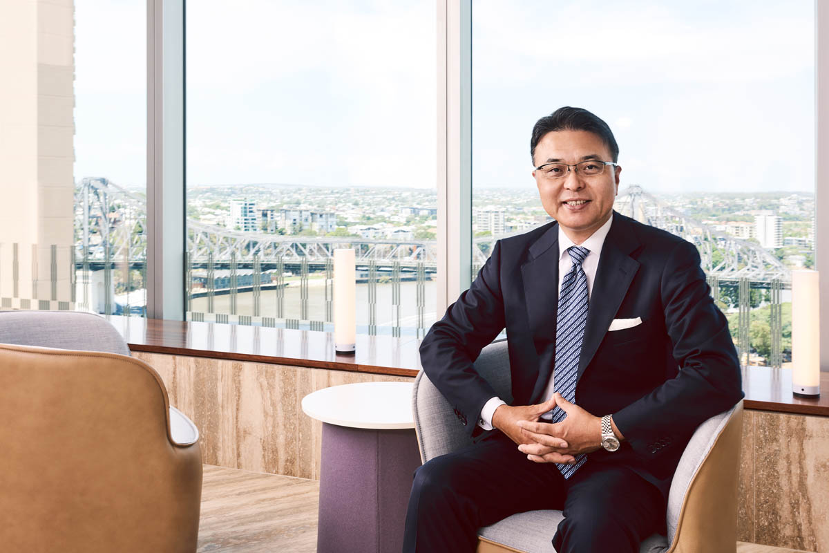 Corporate headshot of man in navy suit and tie, seated in office with city view