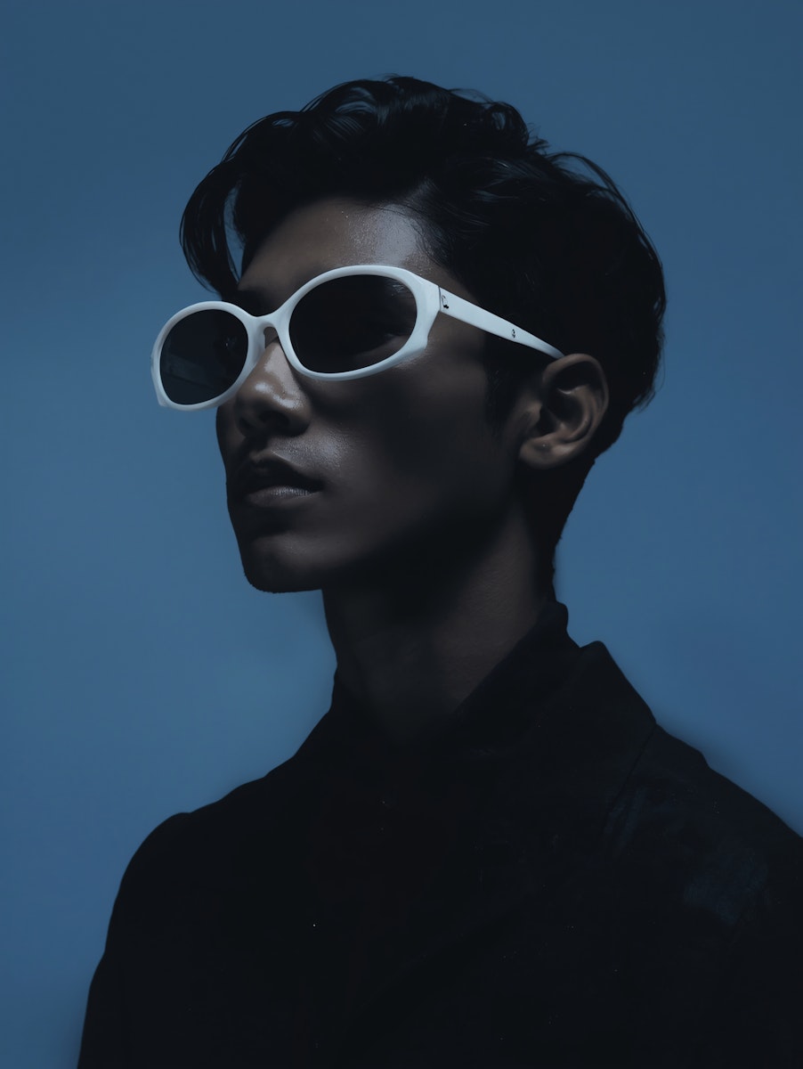 Side-profile portrait of a man wearing white sunglasses against a dark blue background