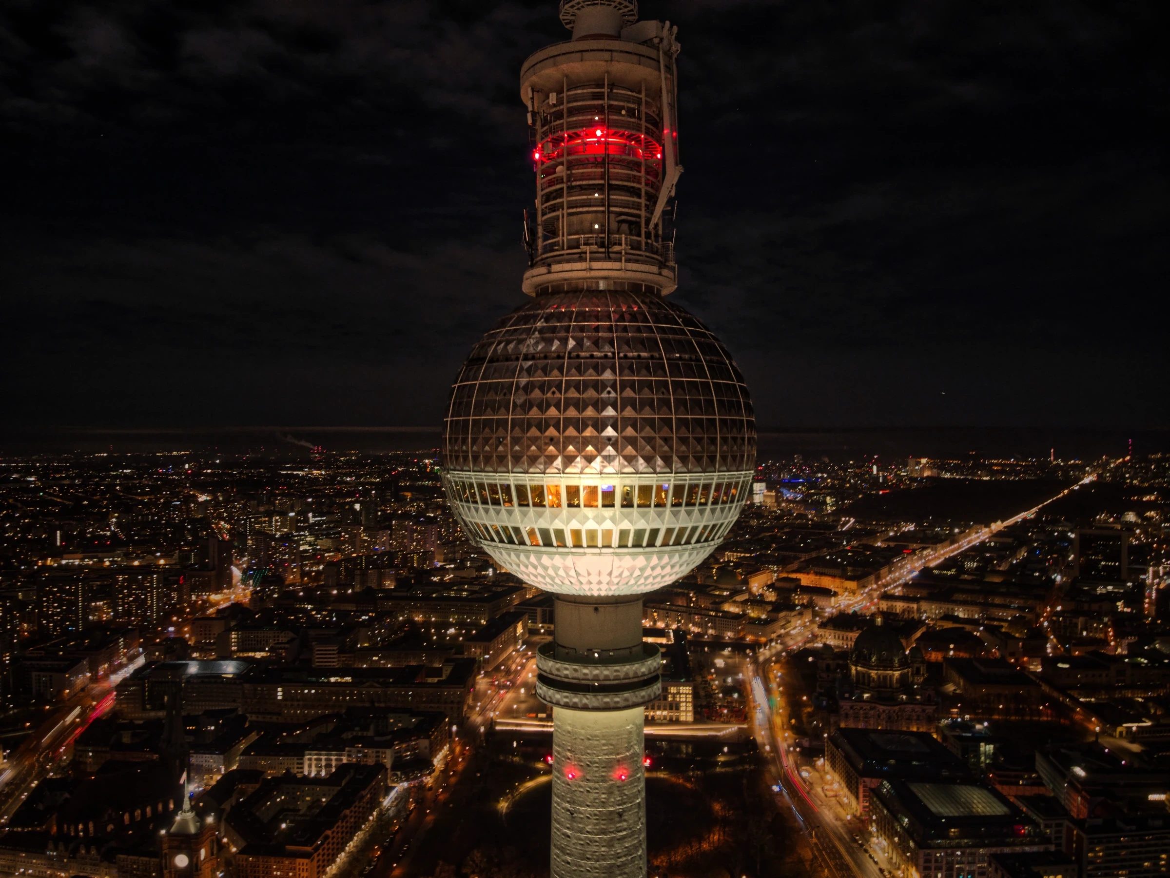 Drohnenaufnahme des Berliner Fernsehturms bei Nacht mit Blick auf die Kugel, das Restaurant Sphere und die beleuchtete Berliner Innenstadt.