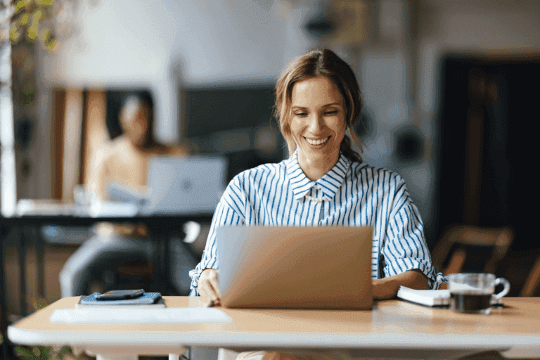 Smiling woman sitting at a desk using a laptop in a bright, modern workspace, with a notebook, phone, and coffee nearby, and a blurred coworker working in the background.