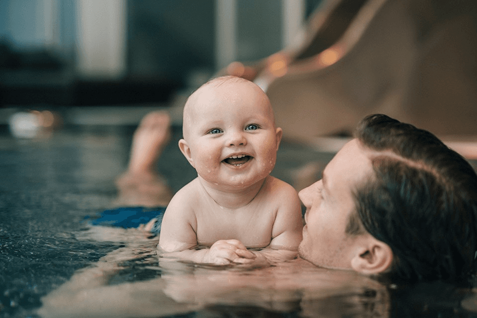 A joyful baby with a big smile is being held by a caregiver in a swimming pool, capturing a heartwarming moment of play and bonding in the water.