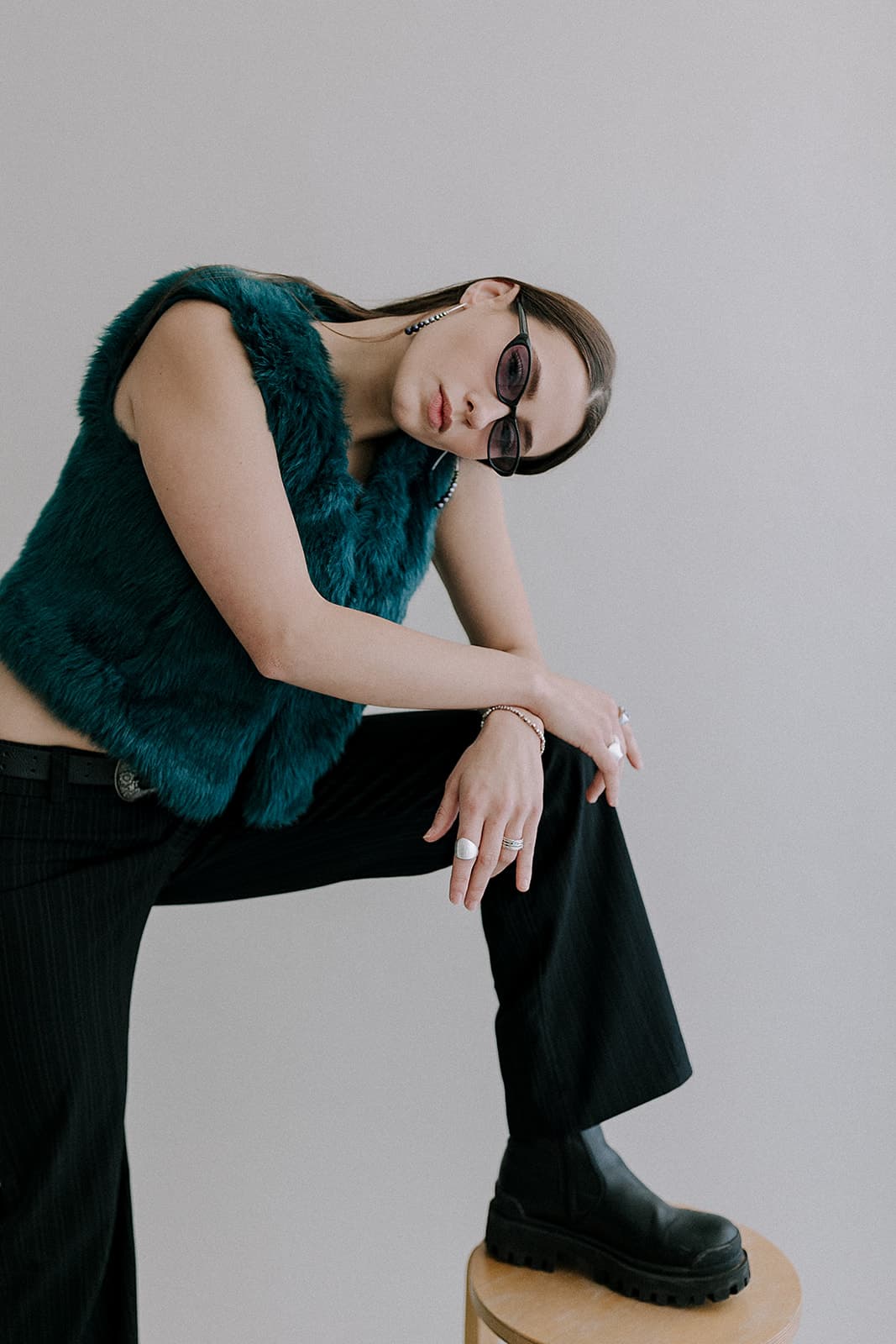 A model leans on a stool wearing a teal fur vest and black sunglasses, photographed in Revelator Studio, a natural light photography studio in Shreveport.