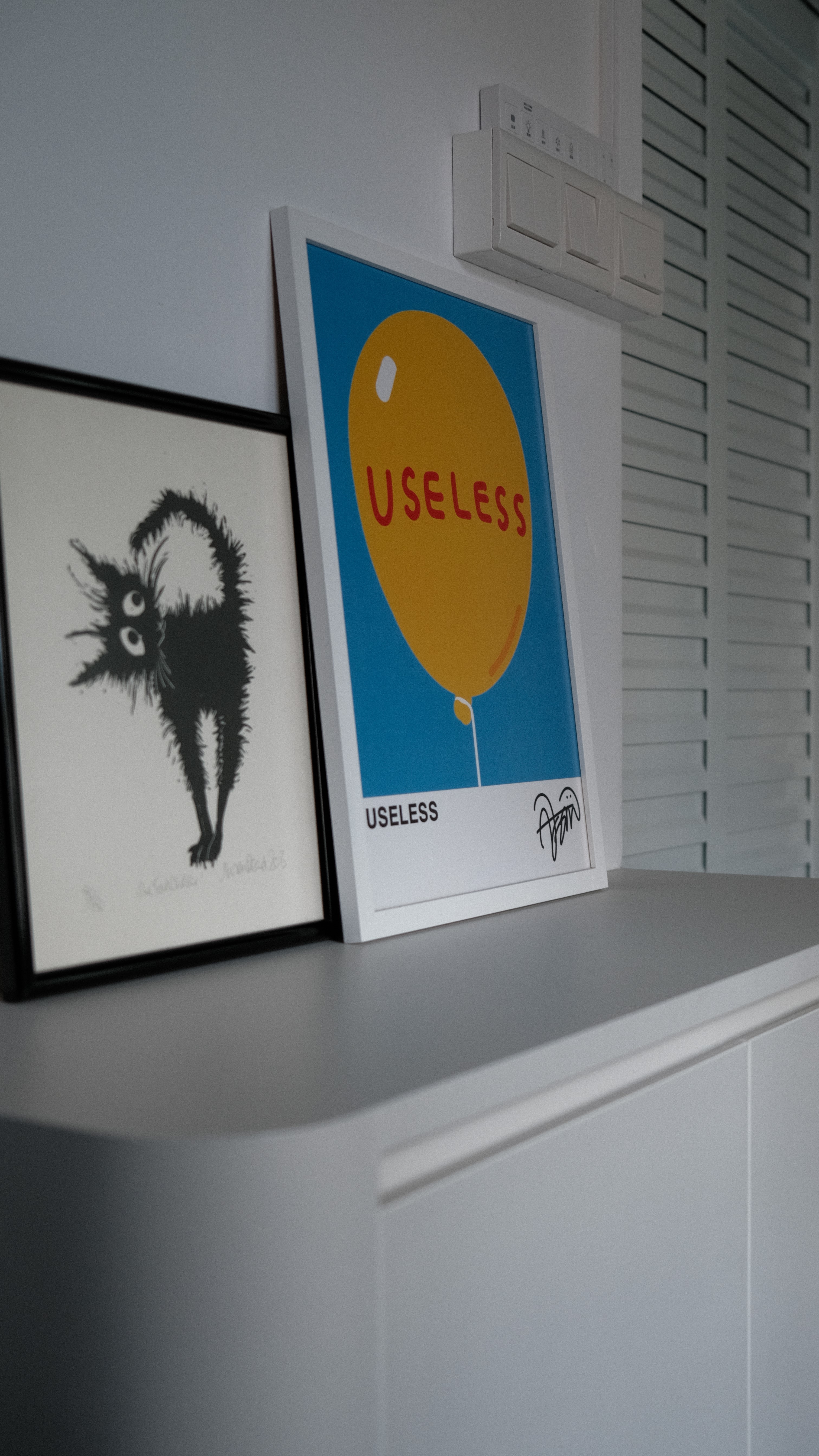 Close-up of framed cat illustration and “Useless” balloon poster atop a minimalist cabinet, emphasizing the home’s quirky art curation in Holland C