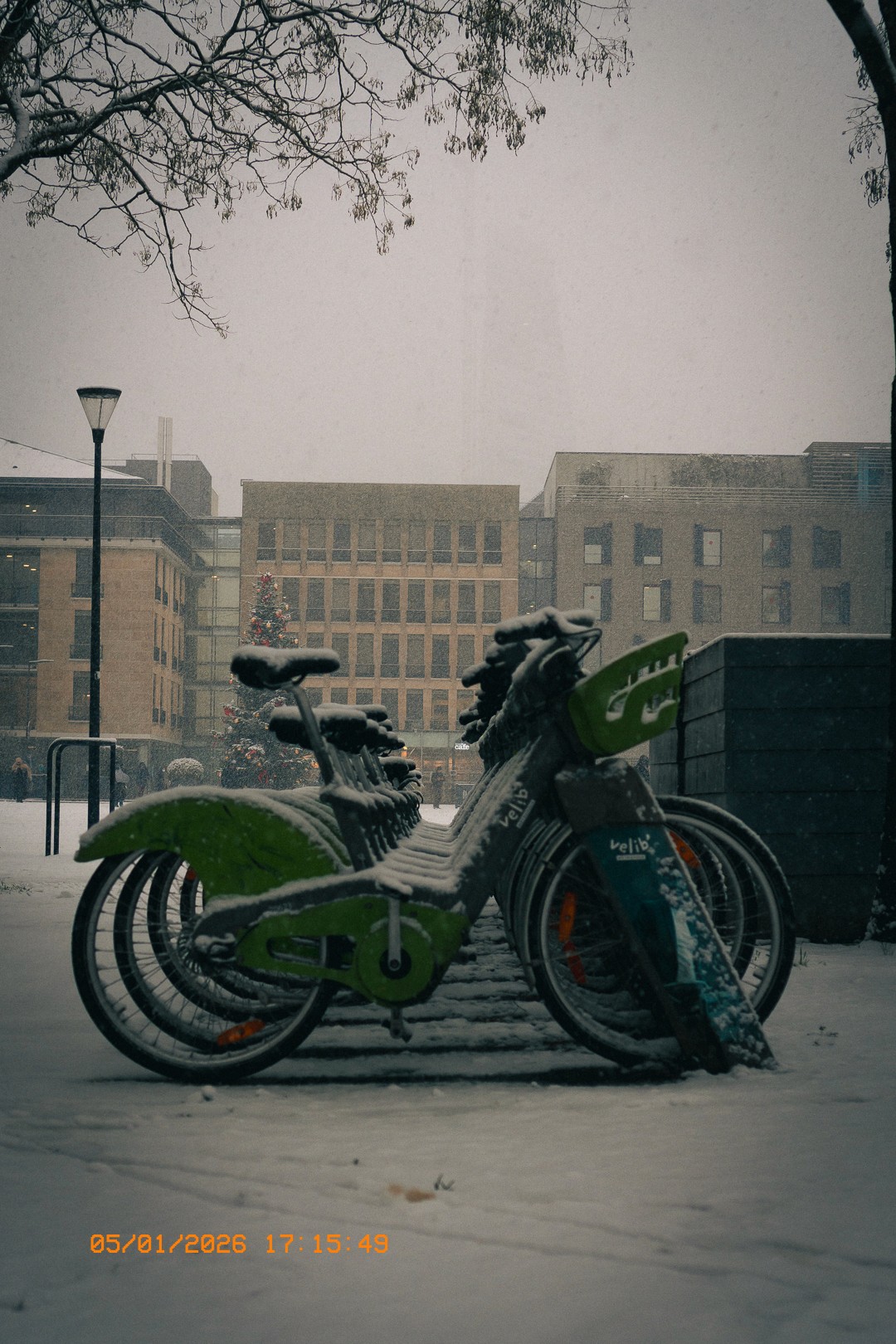 Vélos partagés Velib recouverts de neige dans un paysage urbain hivernal à Issy, avec des immeubles modernes, un arbre de Noël décoré et des branches d'arbres sans feuilles. Ciel gris et neige tombant.