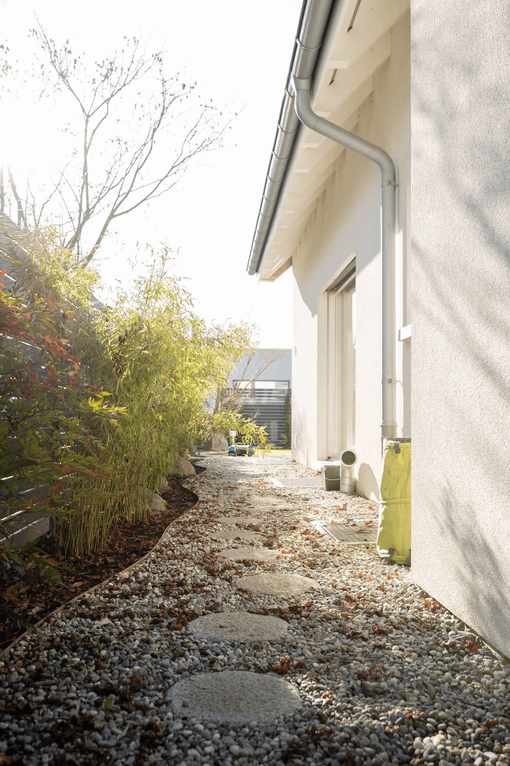 A pathway lined with stones leads to a modern building, surrounded by sparse vegetation and soft sunlight.