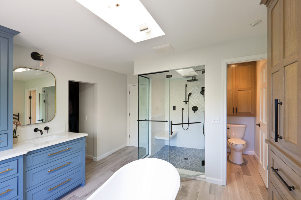 Bright modern bathroom featuring a white freestanding tub, light wood floors, large skylight, and built-in wooden and blue cabinetry. A walk-in glass shower with black fixtures sits near a private toilet room.