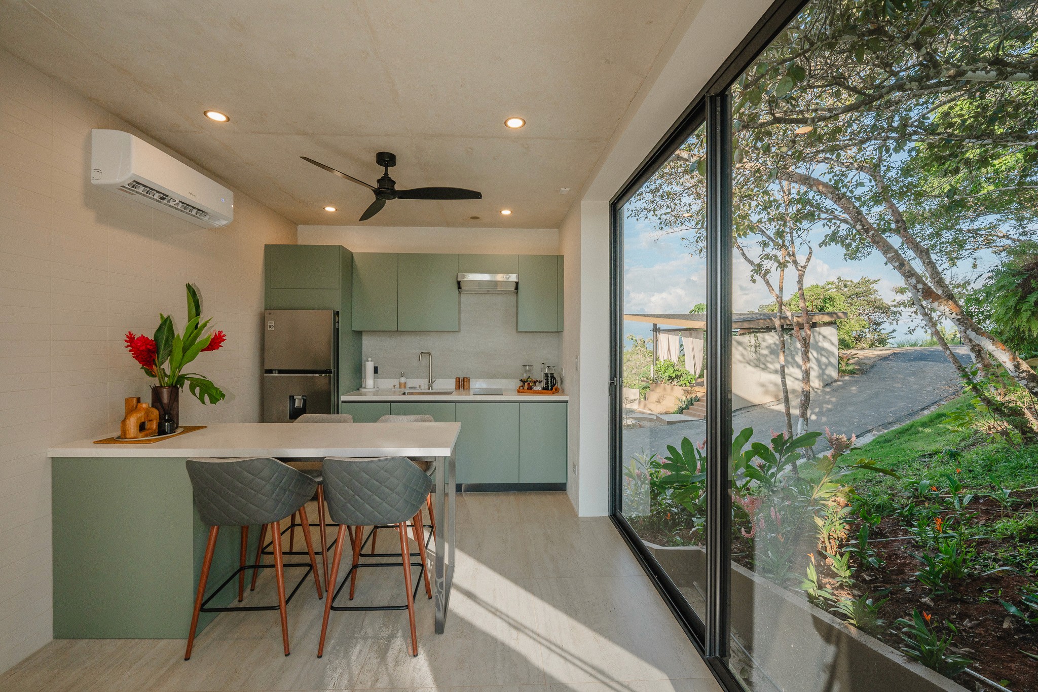 Open-concept kitchen and dining area in a Costa Rica casita. Includes sage green cabinetry, a white breakfast bar with gray stools, and floor-to-ceiling glass doors showcasing the outdoor tropical greenery.