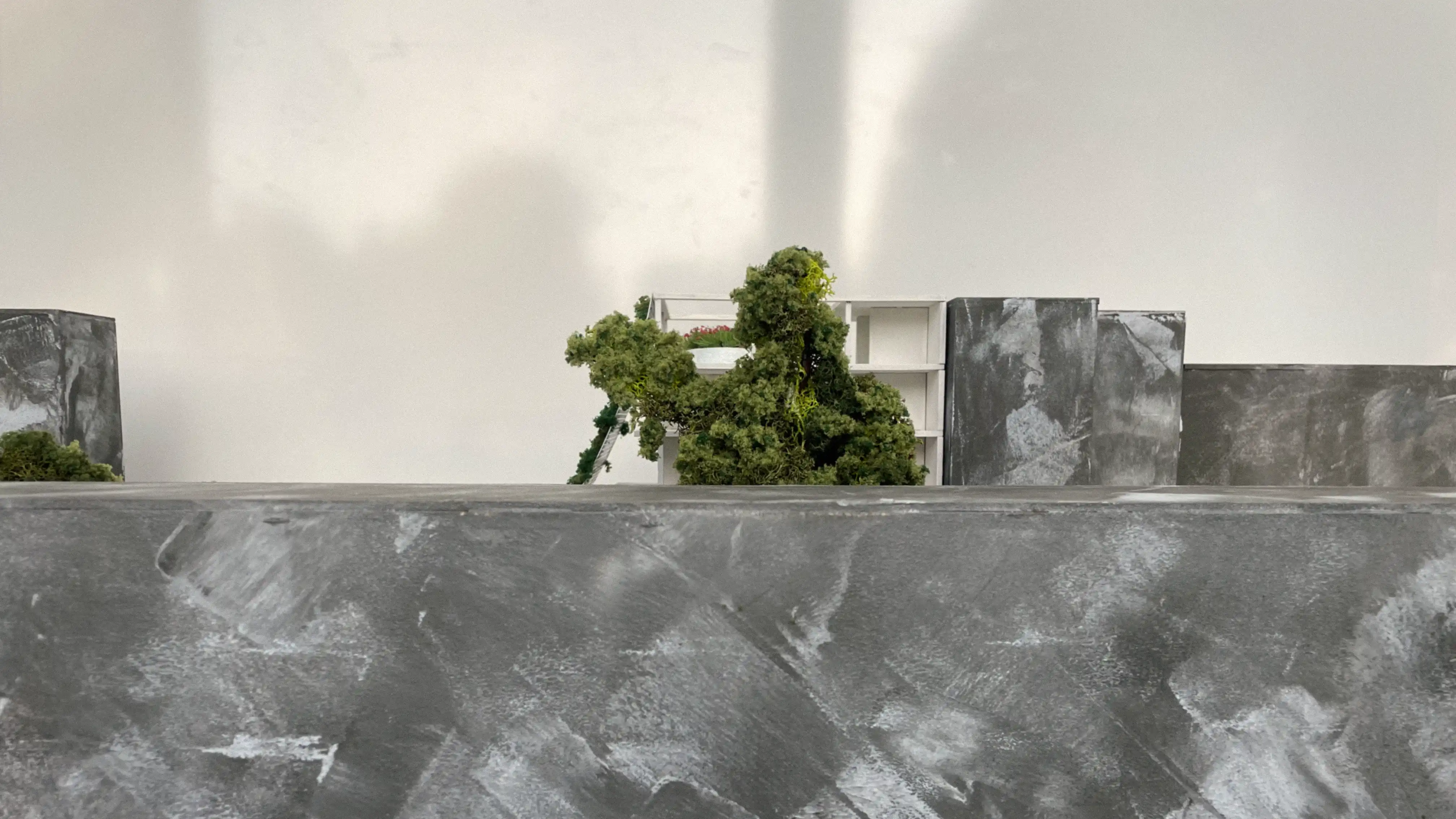 Architekturmodell in Frontalansicht: Nahaufnahme einer geglätteten Sichtbetonmauer im Vordergrund, dahinter ein begrüntes Gebäudeelement mit einer Modell-Leiter und weißer Tragstruktur