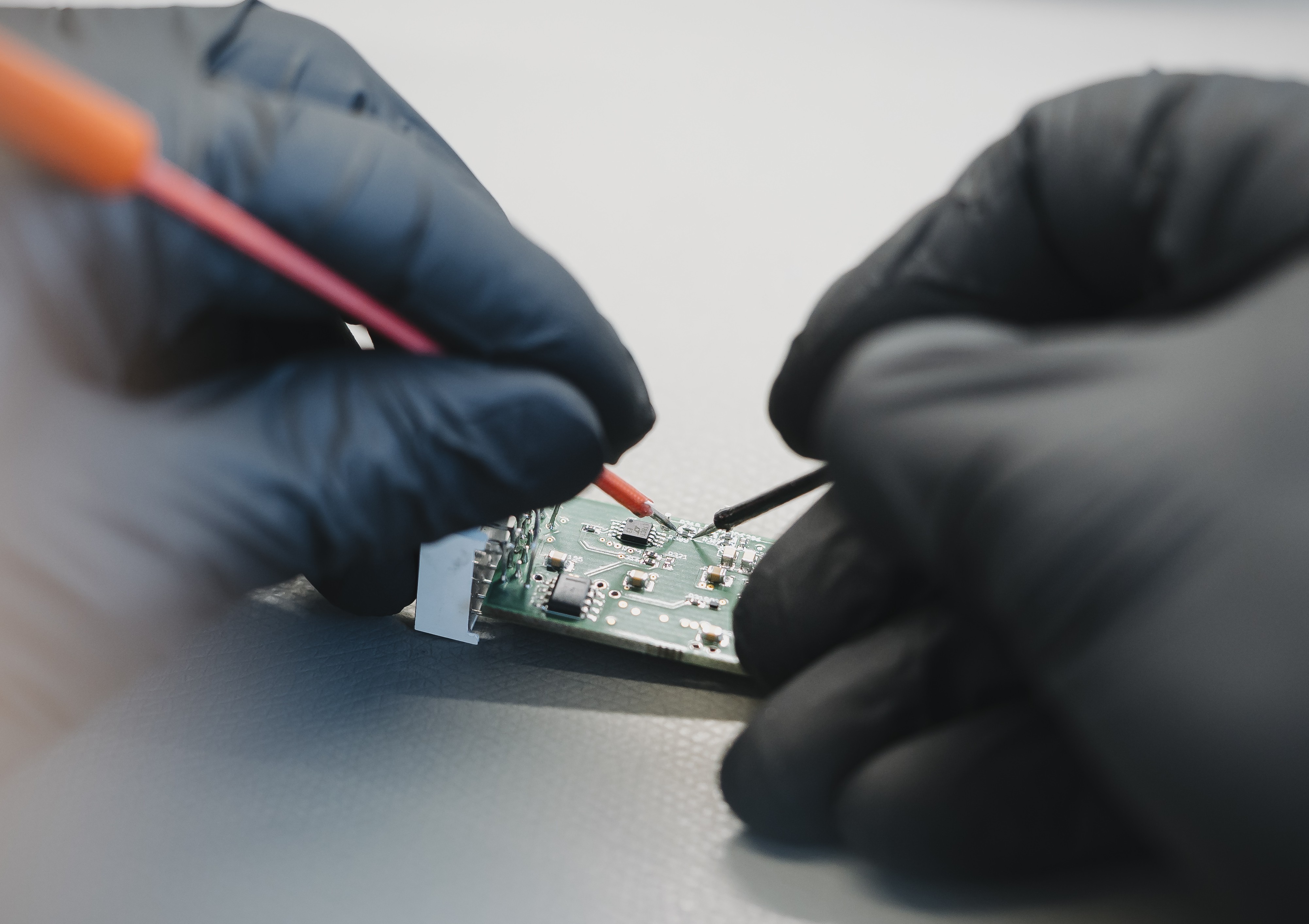 A close-up of two hands in black gloves performing delicate, precise work on a tiny circuit board with a fine tool.