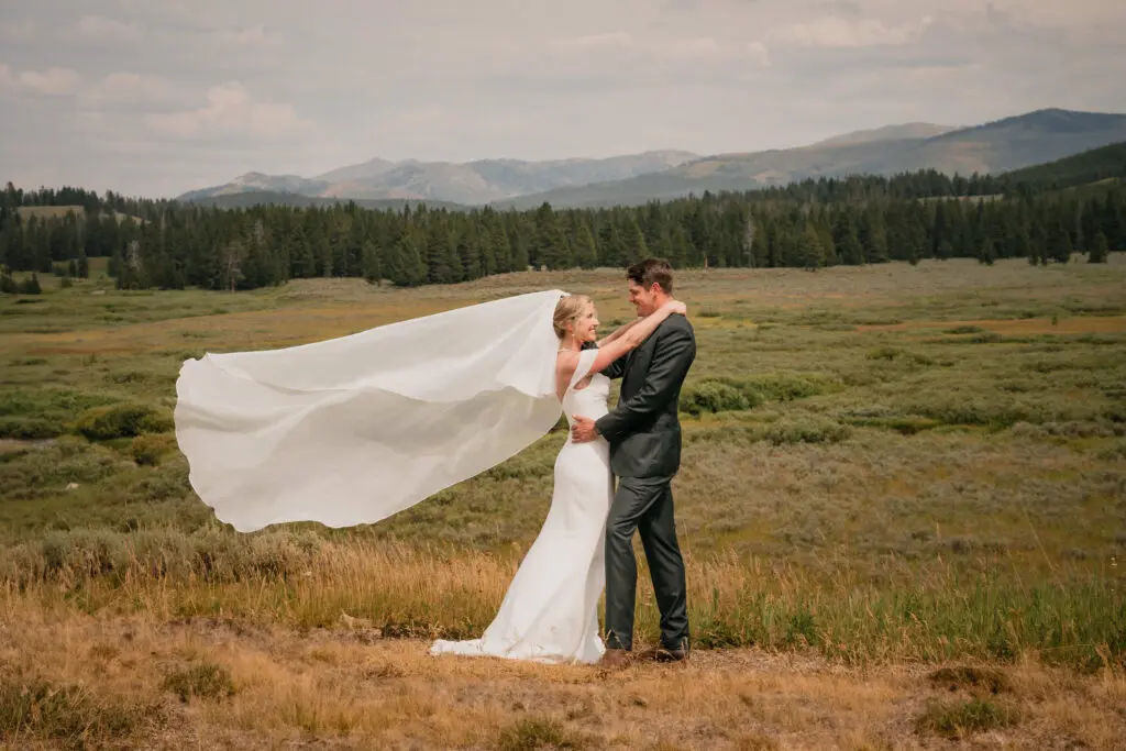 Bride and groom embracing in alpine meadow with flowing white veil catching wind, evergreen forest and mountain range in distance