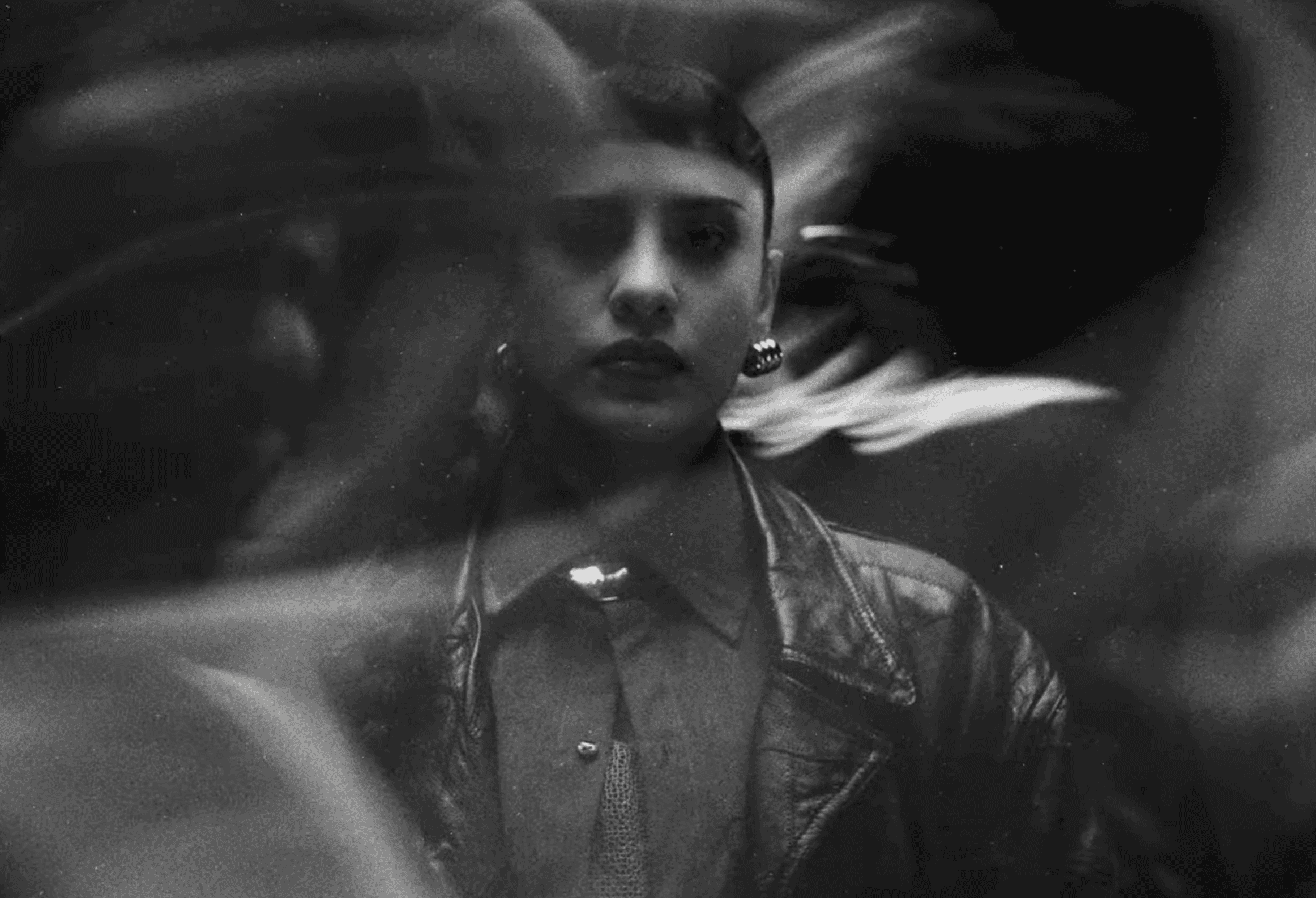Black and white image of GRTSCH with a serious expression, wearing a leather jacket and earrings. Motion blur surrounds them, creating a mysterious tone.