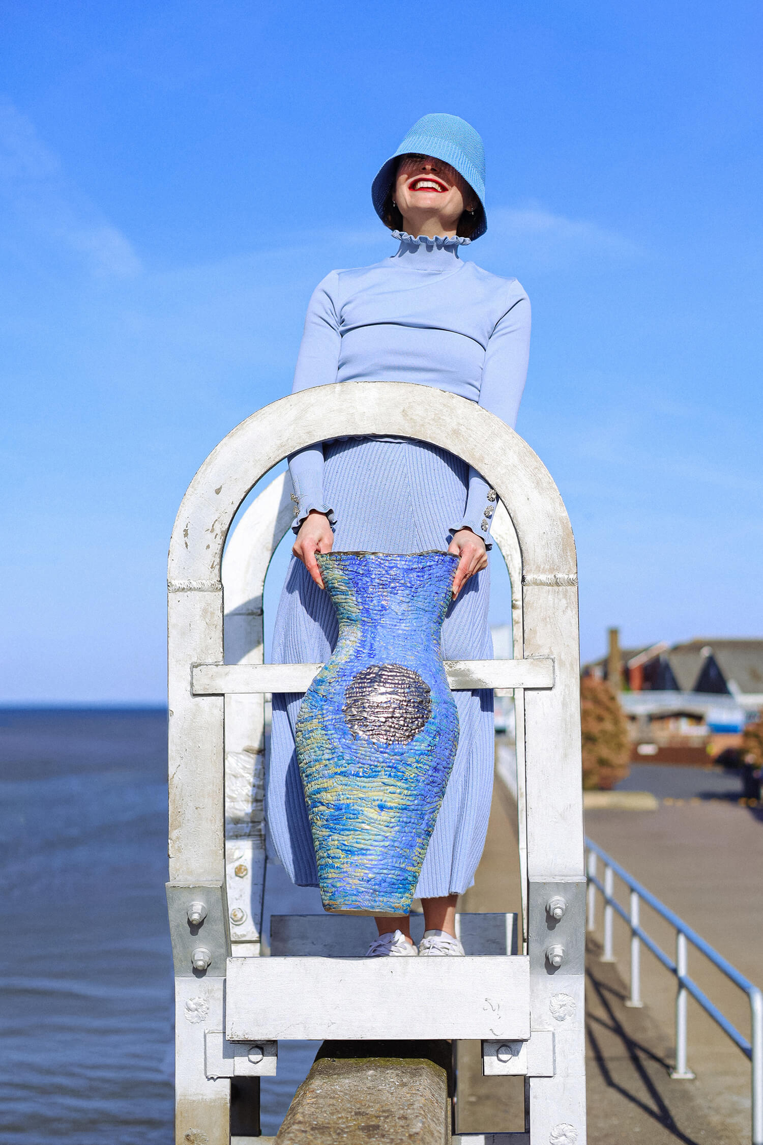 Editorial fashion portrait of woman in pastel blue outfit posing by the seaside with ceramic vase.