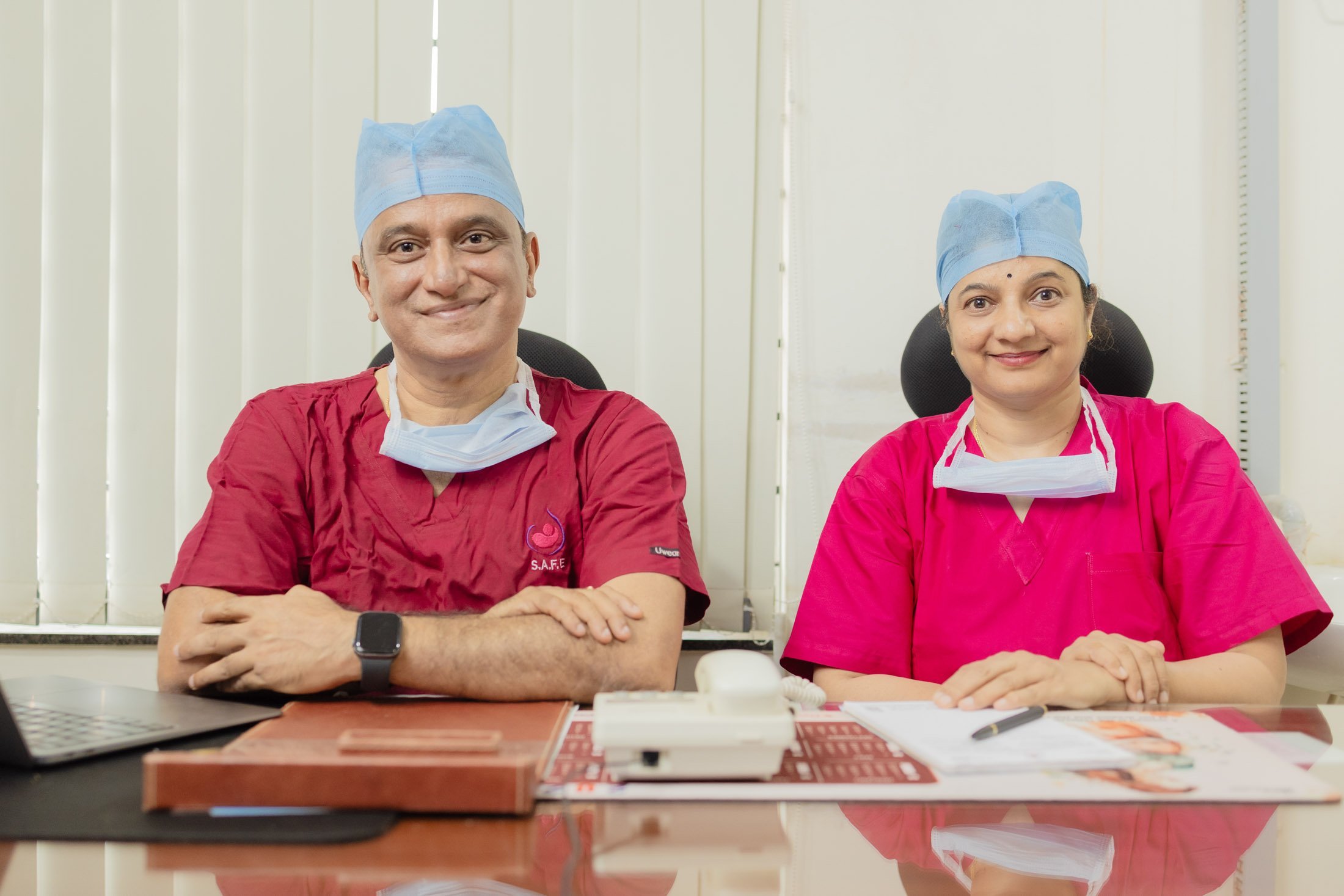 Two doctors in white lab coats and stethoscopes standing confidently in a clinical setting with medical posters and a bed in the background