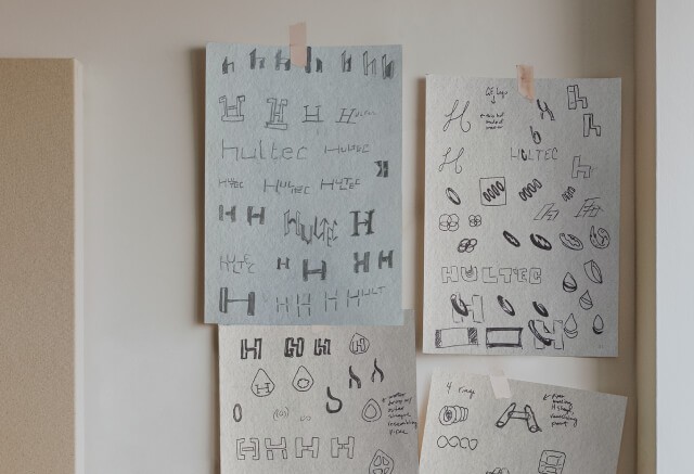 Sheets of paper taped to a wall display various hand-drawn logo sketches and creative design ideas, including different stylized shapes and the word HULTEC, reflecting the brand’s early storytelling process.