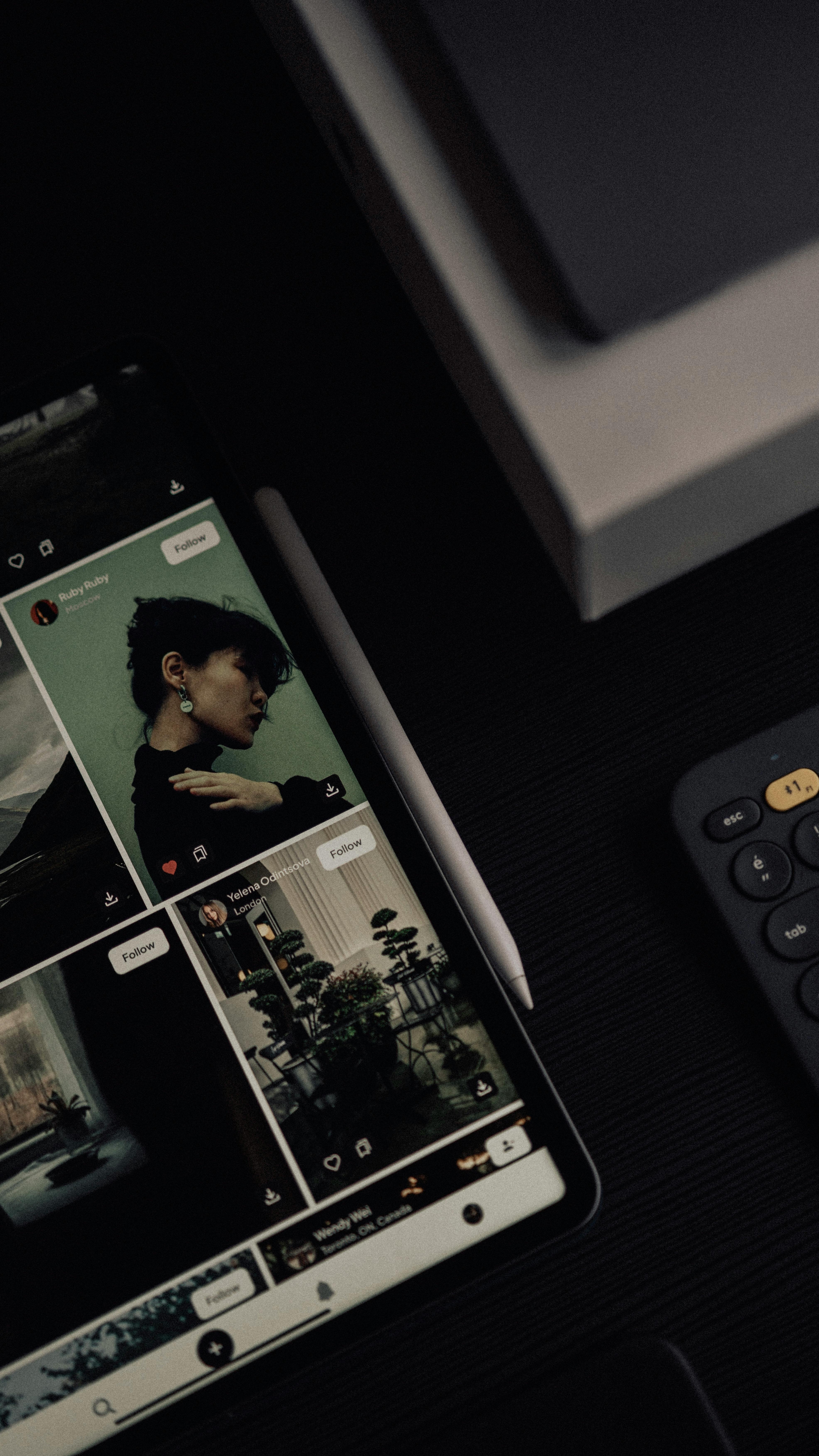Tablet showing a photo feed next to a stylus and keyboard on a dark desk. Moody, creative workspace.