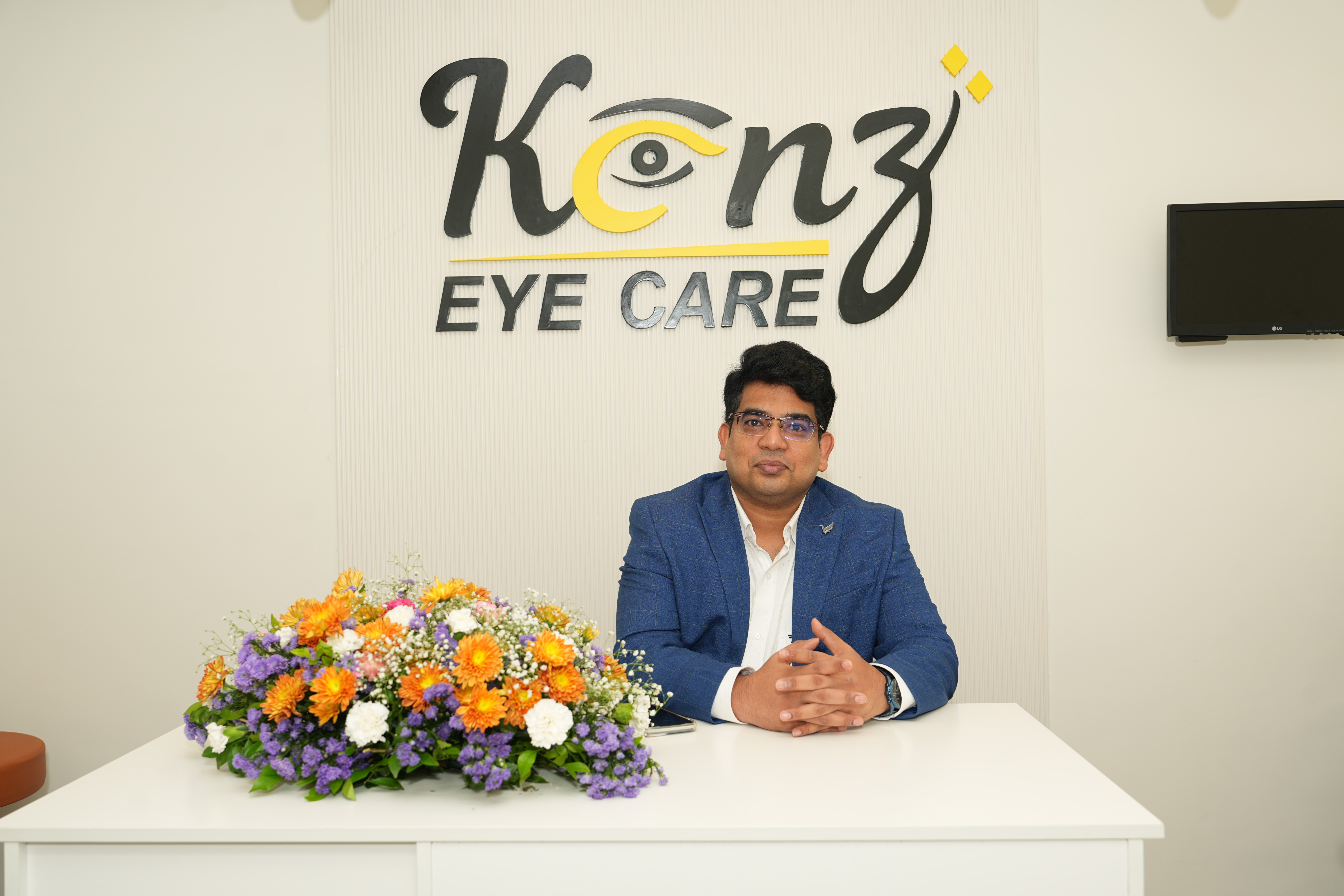 A portrait of Dr. S.M. Qutubuddin Ali, the lead ophthalmologist and retina specialist at Kenz Eye Care in Kokapet, Hyderabad. Dr. Ali is seated professionally at a desk with a vibrant bouquet of flowers, with the clinic's logo clearly visible behind him. This image establishes a personal connection with potential patients and highlights the expert care available at the clinic. It is an ideal SEO asset to attract clients seeking an experienced and trusted eye doctor, retina specialist, or a comprehensive eye check-up in and around the Kokapet area. The image projects professionalism and a welcoming environment, encouraging new clients to schedule an appointment for their eye health needs.