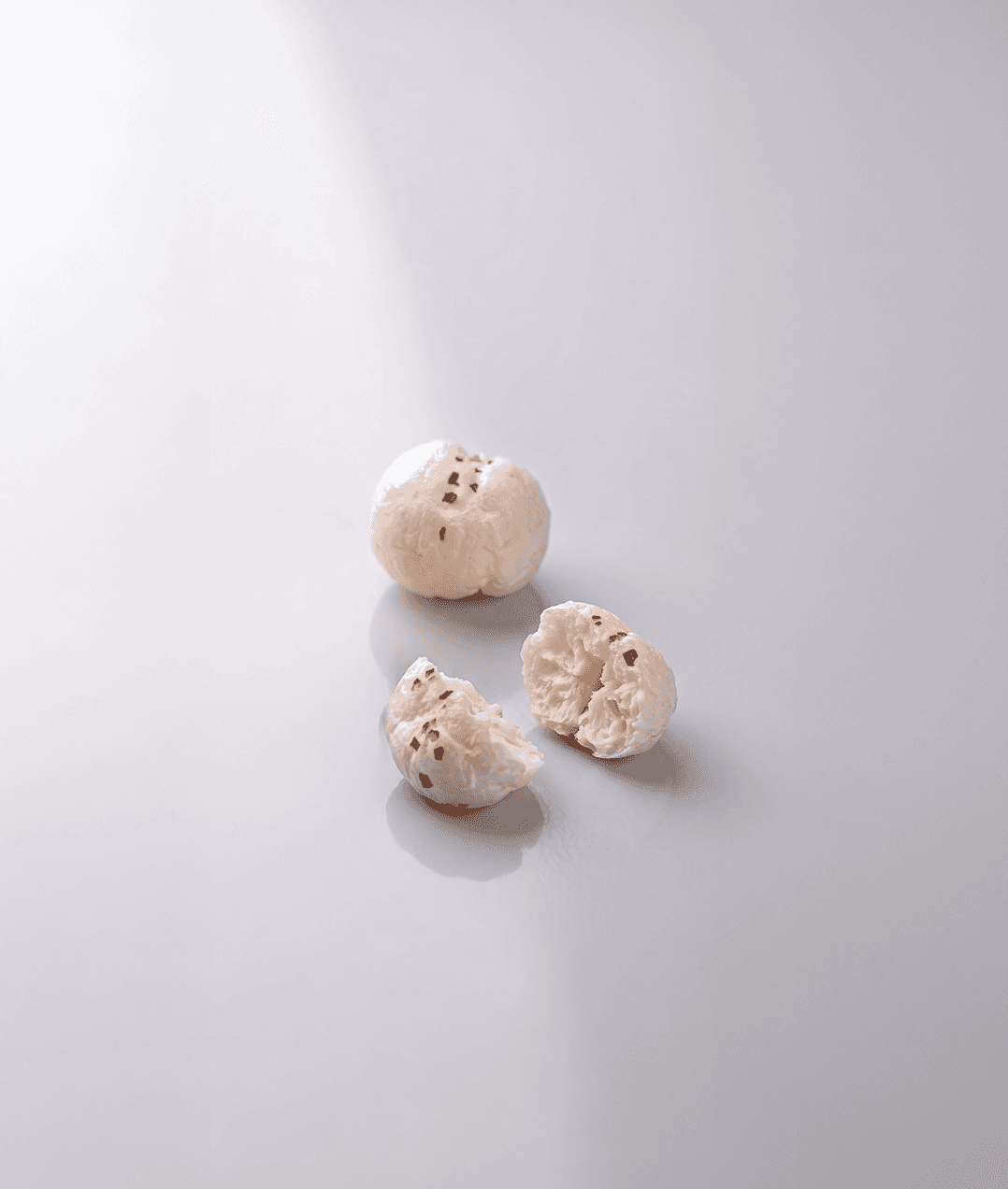 One whole white-coated air-popped lotus seed snack and one broken into two pieces beside it, showing a pale, porous interior with visible herb flecks, on a plain light grey surface.