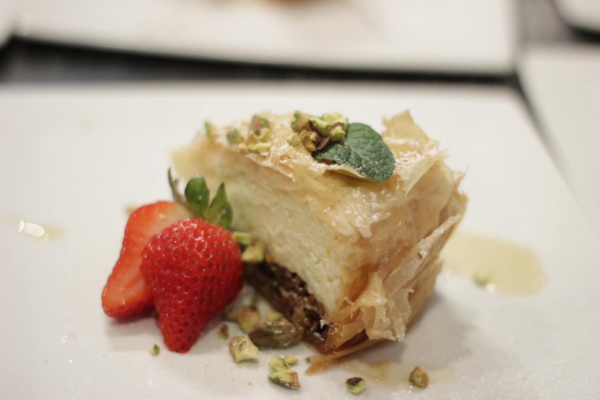 Plated Pistachio Baklava Cheesecake slice with crispy golden phyllo layers, topped with crushed pistachios and a fresh mint leaf, served alongside sliced strawberries on a white plate.