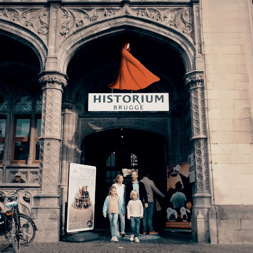 Egy négytagú család áll a Historium Brugge bejáratánál, egy történelmi épület díszes kőívekkel és feliratokkal.