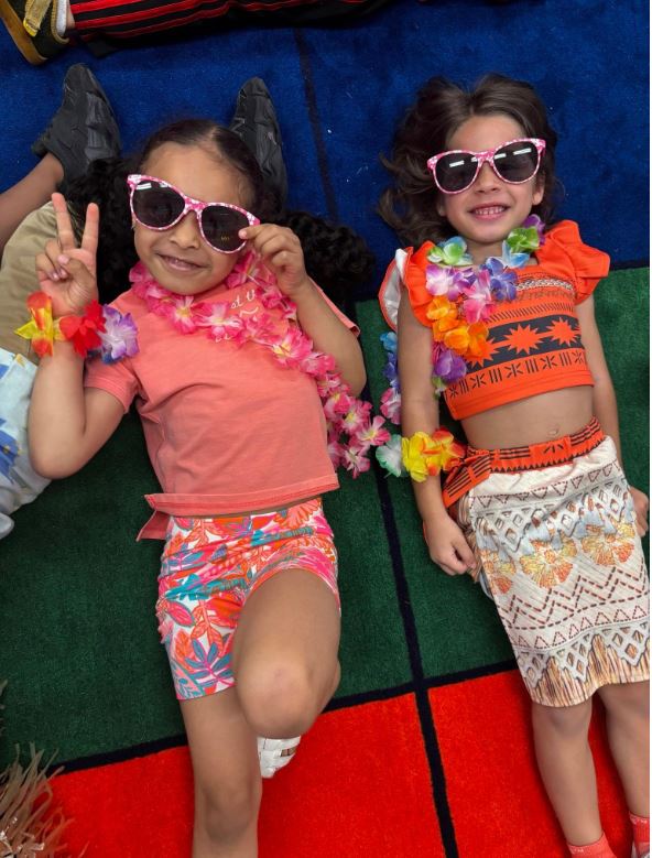 Two children pretend to lay on the beach donning sunglasses, leis, and other beachwear.