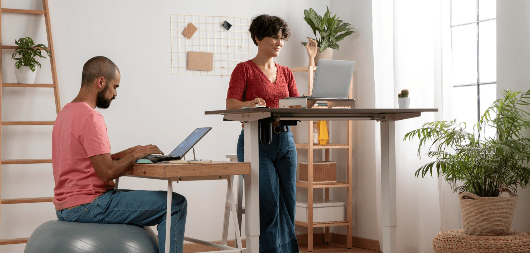 Two people working in a bright home office — one sitting on an exercise ball at a desk, the other standing at a height-adjustable desk, surrounded by plants and natural light.