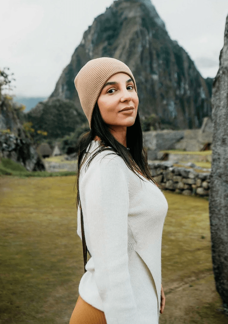 A smiling woman with her arms crossed, standing against a dark green background. She has long, dark hair.