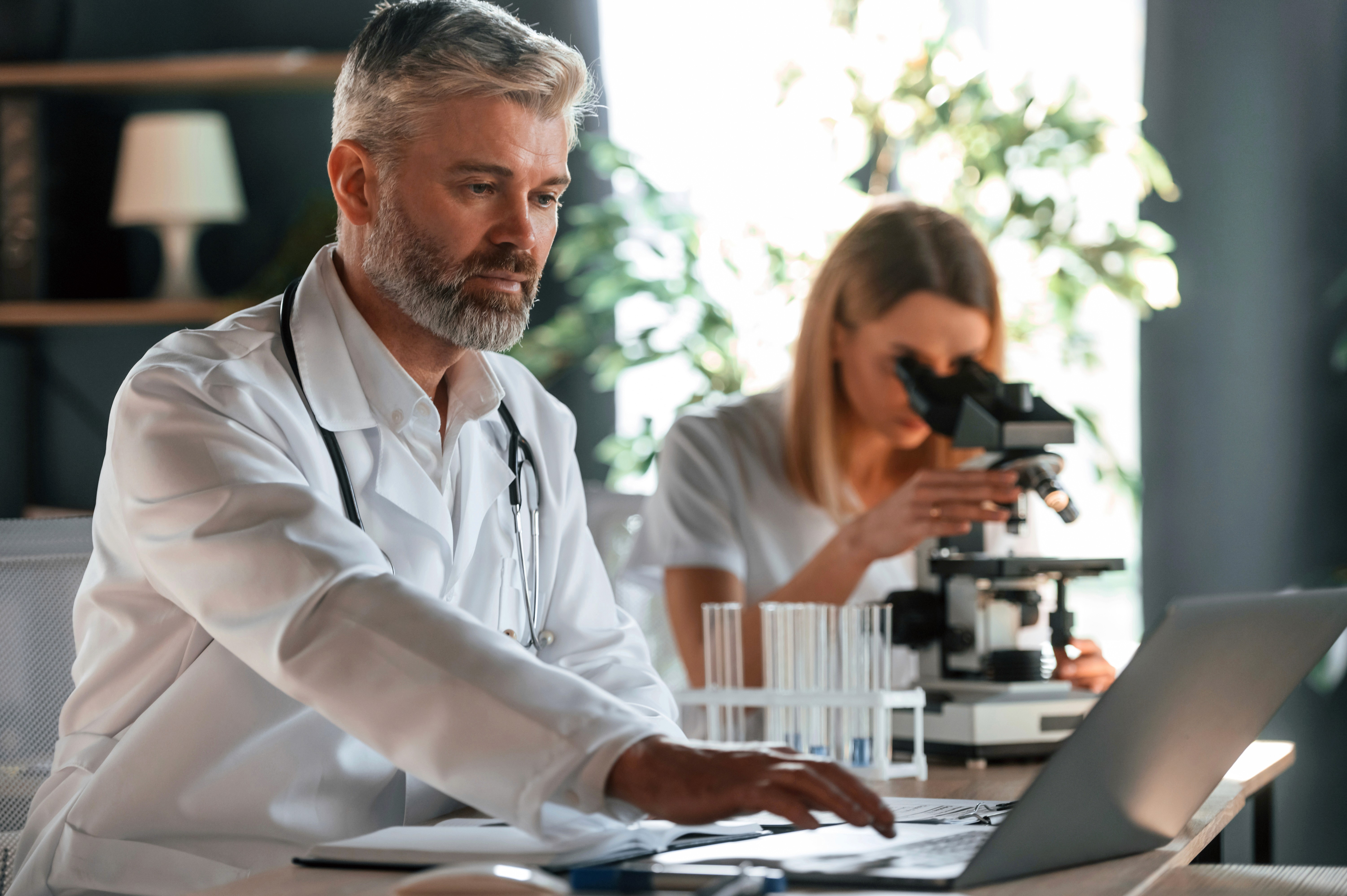 A man in a white lab coat working at a computer, viewing a 3D model on the screen in a laboratory or office setting.