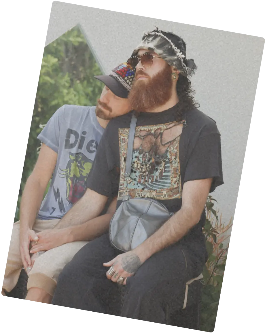 Two bearded people sitting together, one resting their head on the other’s shoulder in a quiet, affectionate moment.
