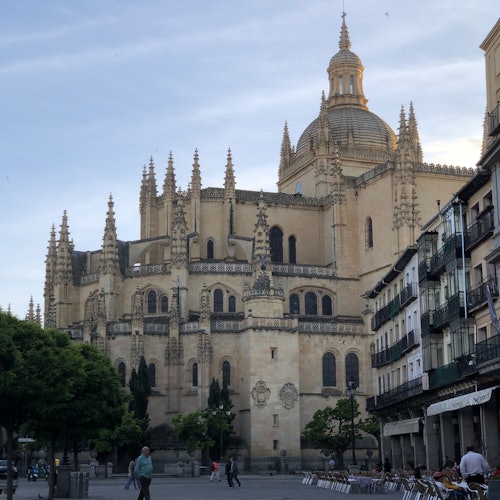 A cathedral with elaborate spires and a dome, positioned in a square with people, trees, and surrounding buildings.