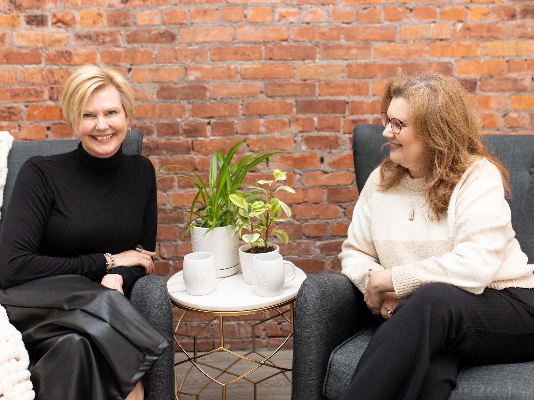 Image of Two women sitting on gray chairs with plants and coffee cups on a small white table.