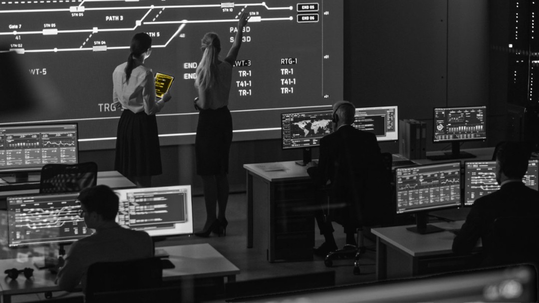 Black and white photo of a cyber analysis room. At the screen in front are 2 woman researchers analysing what is on screen. 3 other male researchers are working on computers facing the big screen.