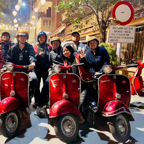 Gruppe mennesker med hjelme poserer med røde scootere på en lyst oplyst gade om natten.