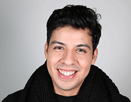 The image shows a man with dark hair wearing a black scarf and a dark-colored shirt. He is smiling and looking directly at the camera against a light gray background.