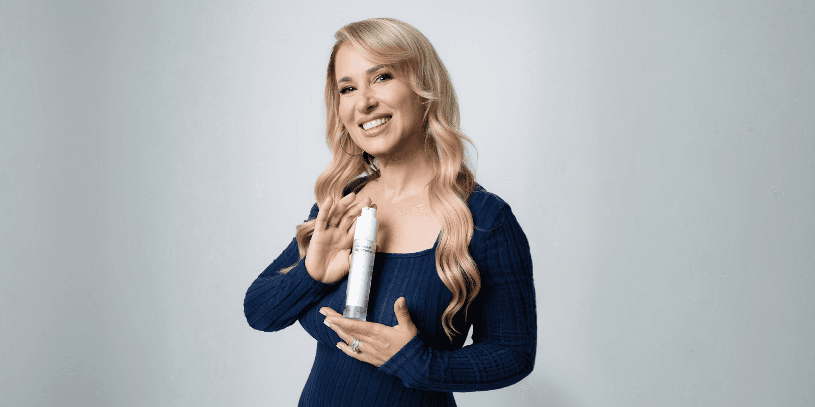 Smiling woman with wavy blonde hair holds a skincare bottle, representing Purely Bella Skincare's mission to empower women.
