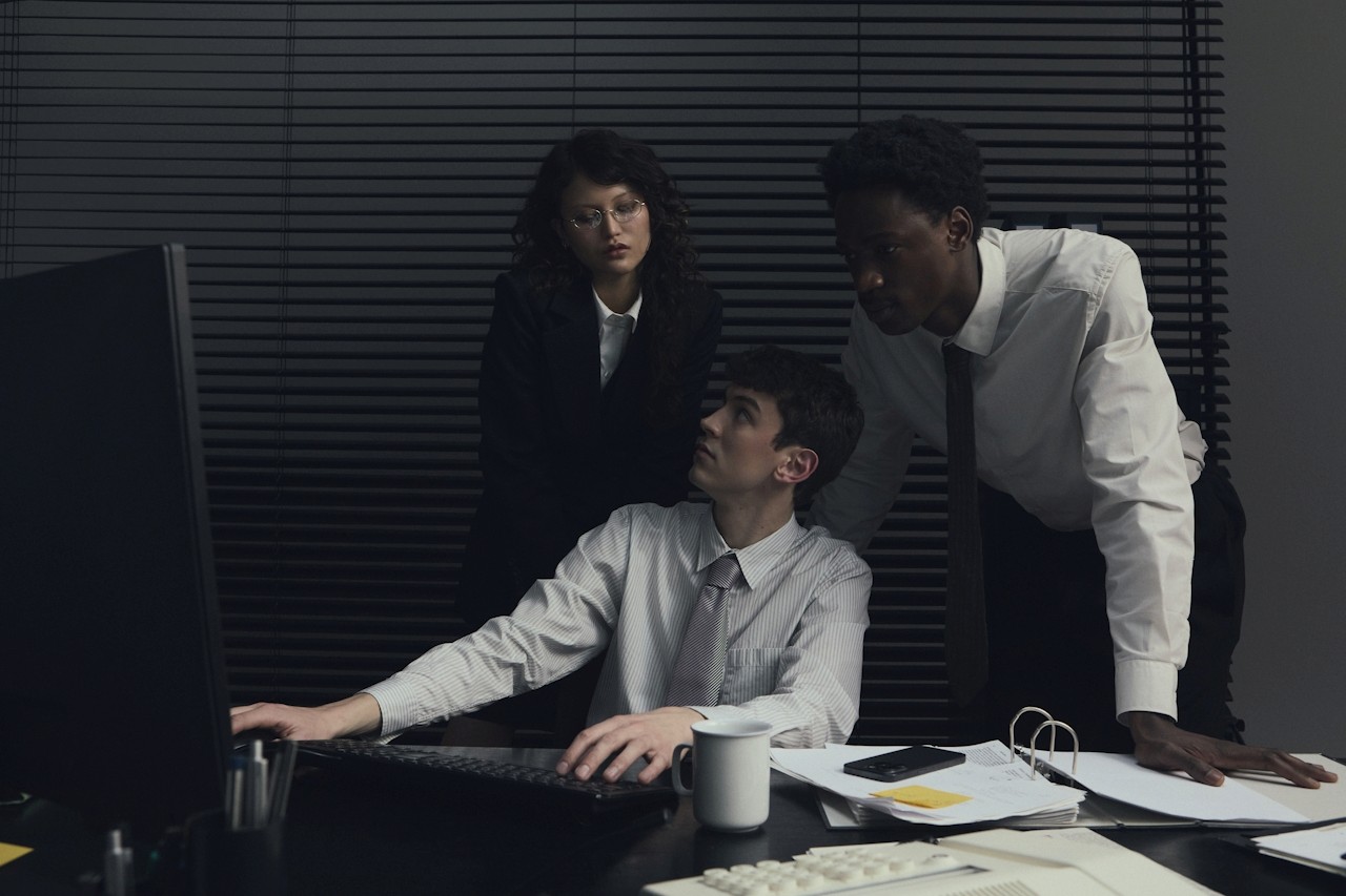 Three colleagues in a dimly lit office look at a desktop computer screen together.