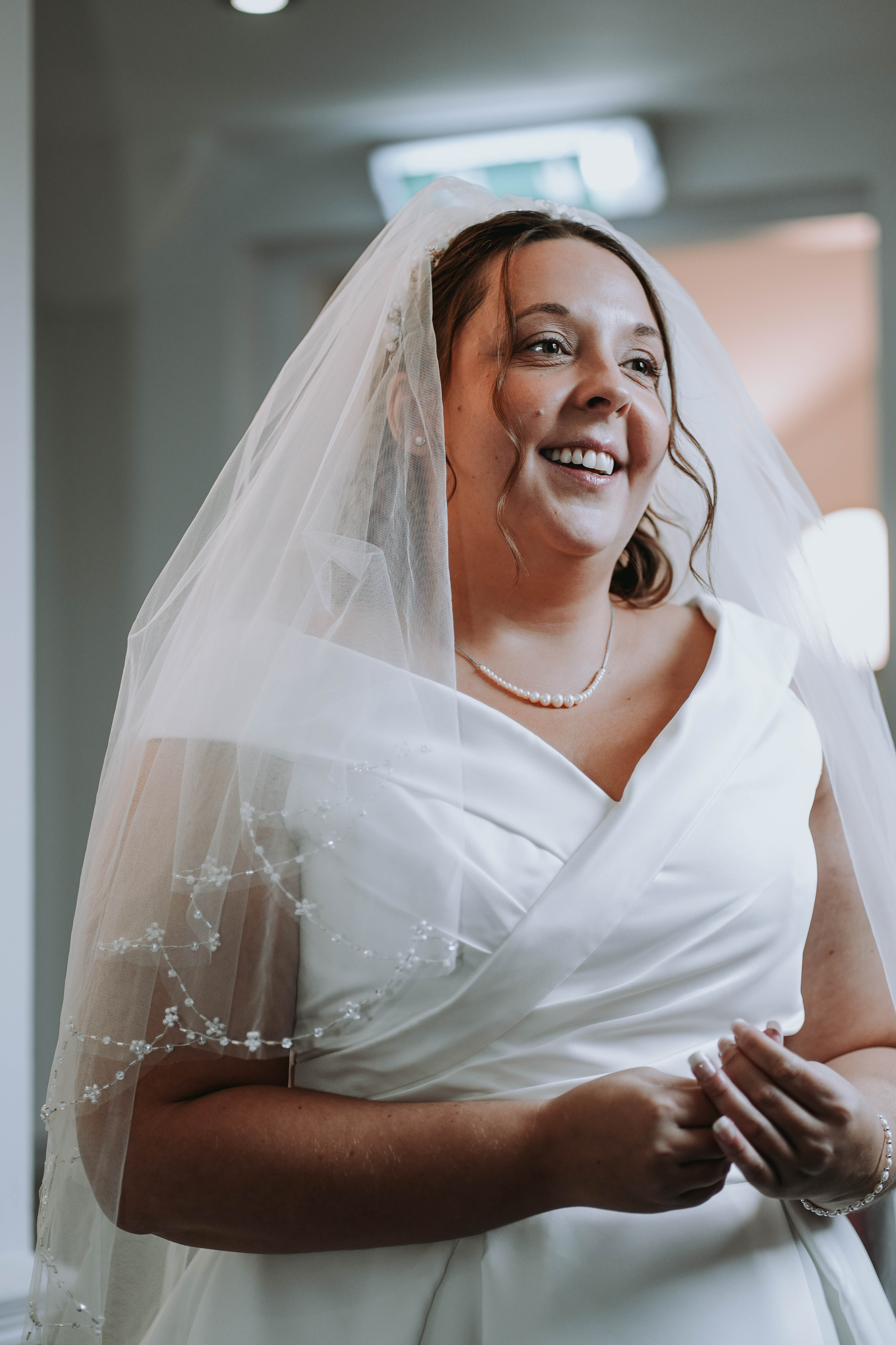 A joyful, candid photo of the bride smiling warmly. She is wearing her white wedding gown, a pearl necklace, and a veil with delicate embroidery on the edge.