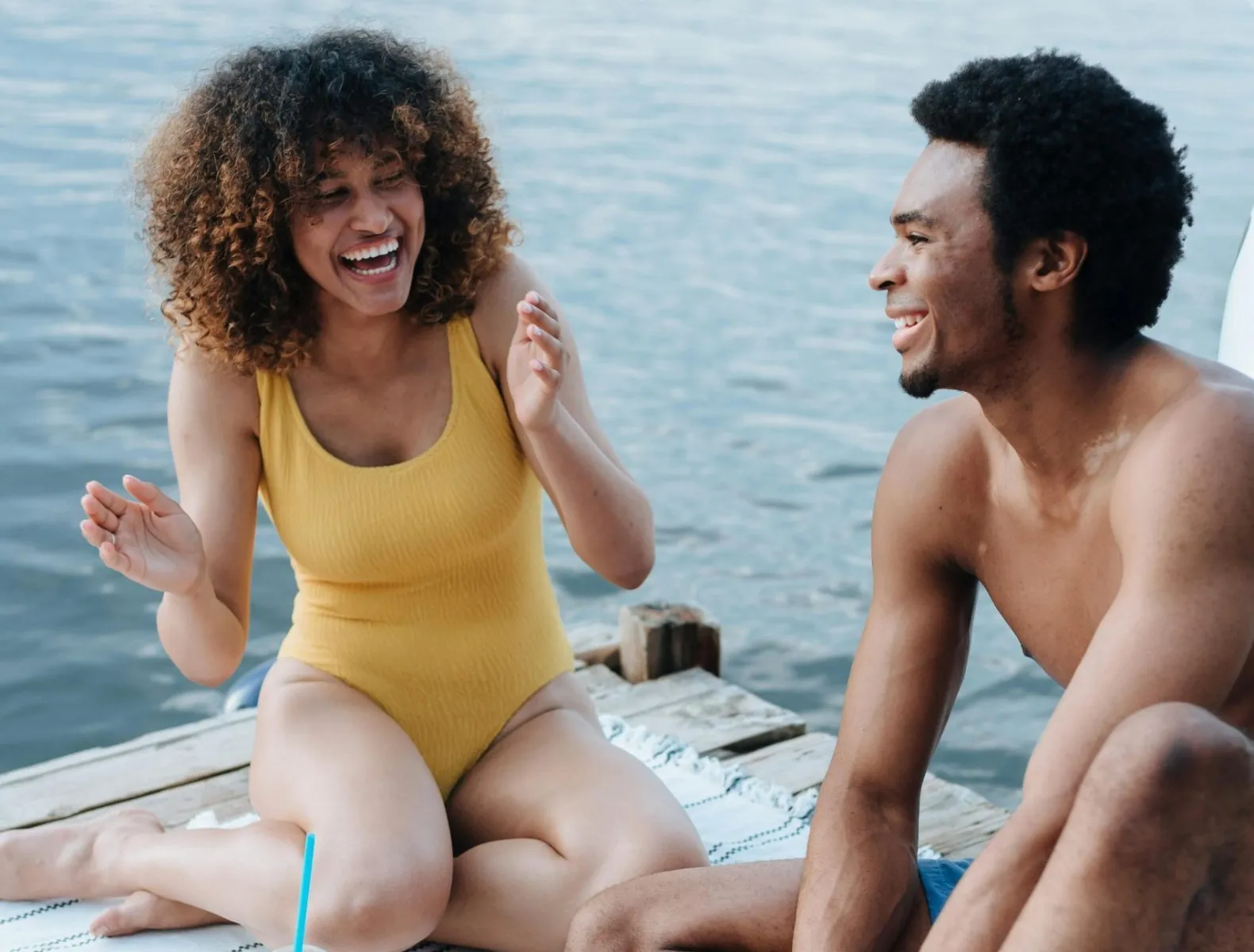 A couple in their bathing suits laughing on a dock at a lake. 