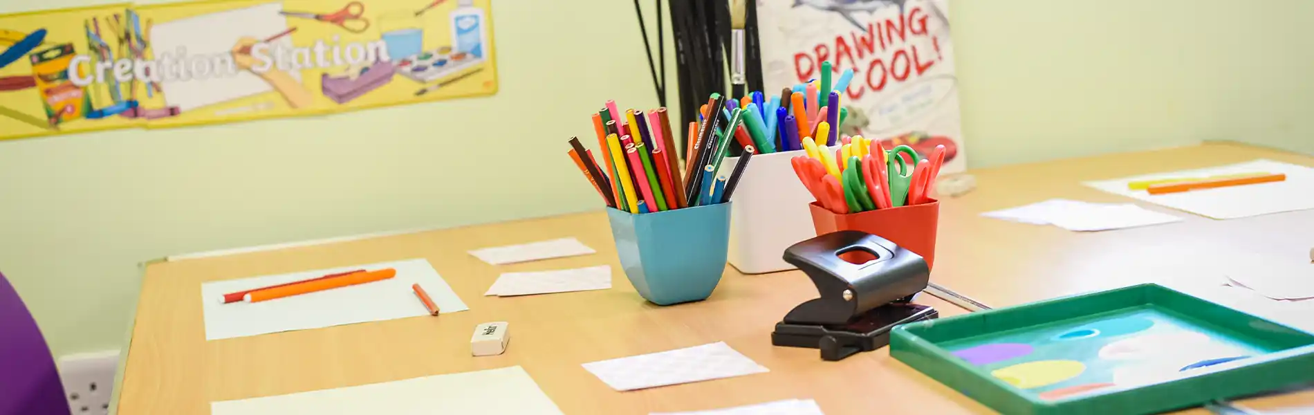 Art supplies on a wooden table: colored pencils, markers, a hole punch, paper, and a green tray. "Drawing Cool!" poster in background.